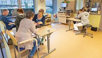 Großer Therapieraum mit mehreren Arbeitsplätzen: Eine Therapeutin begleitet eine erwachsene Patientin bei einer feinmotorischen Übung, während im selben Raum weitere Erwachsene und ein Kind an separaten Tischen therapeutische Aufgaben bearbeiten.