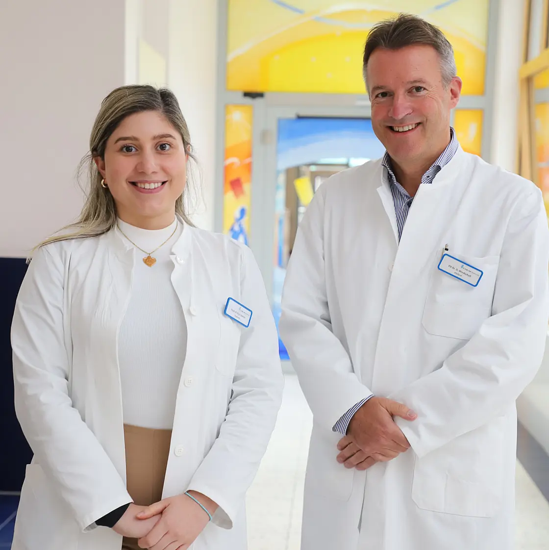 An international physician and a senior specialist stand in a hospital corridor; the image represents the scholarship program offering hands-on training in joint injections, musculoskeletal ultrasound, management of complex paediatric rheumatologic diseases and participation in research projects.