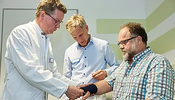 Dr. med. Ansgar Platte und ein Mitarbeiter der Orthopädischen Werkstatt passen gemeinsam eine Bandage am Unterarm eines Patienten an.