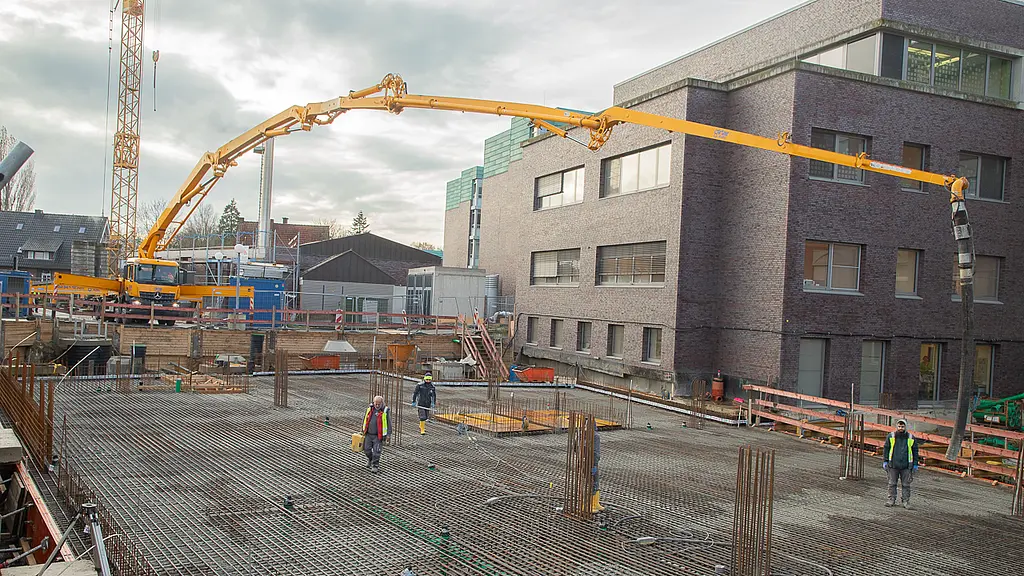 Weitwinkelansicht der Baustelle mit großflächiger Stahlbewehrung. Ein Betonpumpenfahrzeug mit ausgefahrenem Arm steht neben dem Bestandsgebäude und fördert Beton in die Baugrube.