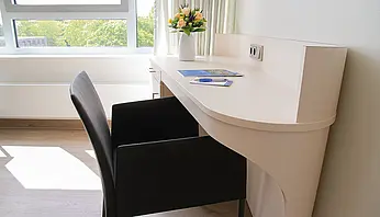 Schreibtisch mit Stuhl am Fenster, darauf Informationsmaterial und ein Blumenstrauß.