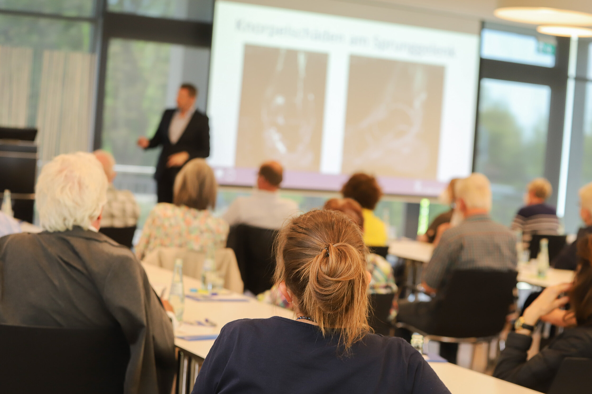 Teilnehmende sitzen an Tischen und hören einem Vortrag zu; im Vordergrund sind Personen von hinten zu sehen, im Hintergrund steht ein Referent vor einer Präsentationsfolie in einem hellen Veranstaltungsraum.