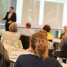 Teilnehmende sitzen an Tischen und hören einem Vortrag zu; im Vordergrund sind Personen von hinten zu sehen, im Hintergrund steht ein Referent vor einer Präsentationsfolie in einem hellen Veranstaltungsraum.