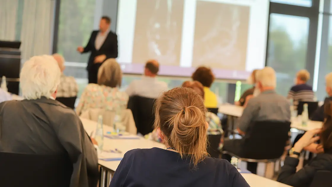 Teilnehmende sitzen an Tischen und hören einem Vortrag zu; im Vordergrund sind Personen von hinten zu sehen, im Hintergrund steht ein Referent vor einer Präsentationsfolie in einem hellen Veranstaltungsraum.