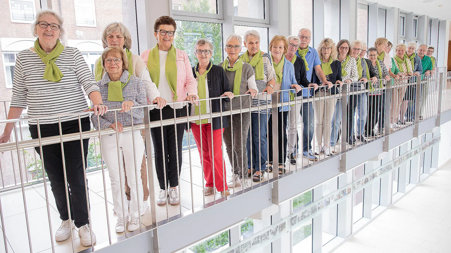 Mehrere ehrenamtliche Krankenhaushilfen stehen nebeneinander an einem Geländer in einem lichtdurchfluteten Flur.