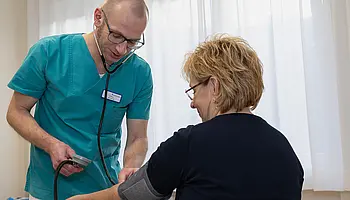 Ein Pfleger mit Stethoskop misst den Blutdruck einer Patientin, die auf einem Stuhl sitzt.