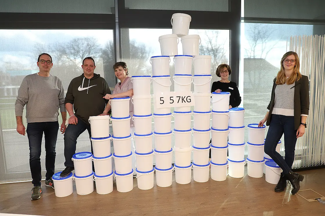 Mitarbeitende stehen neben gestapelten Mehrwegbehältern; ein Schild weist auf die gesammelte Menge eingesparter Verpackung hin.