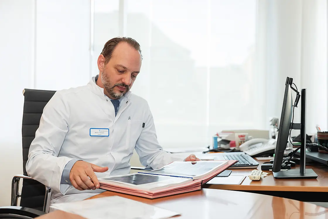 Chefarzt PD Dr. med. Peter Korsten sitzt an seinem Schreibtisch und sichtet medizinische Unterlagen in der rheumatologischen Klinik.