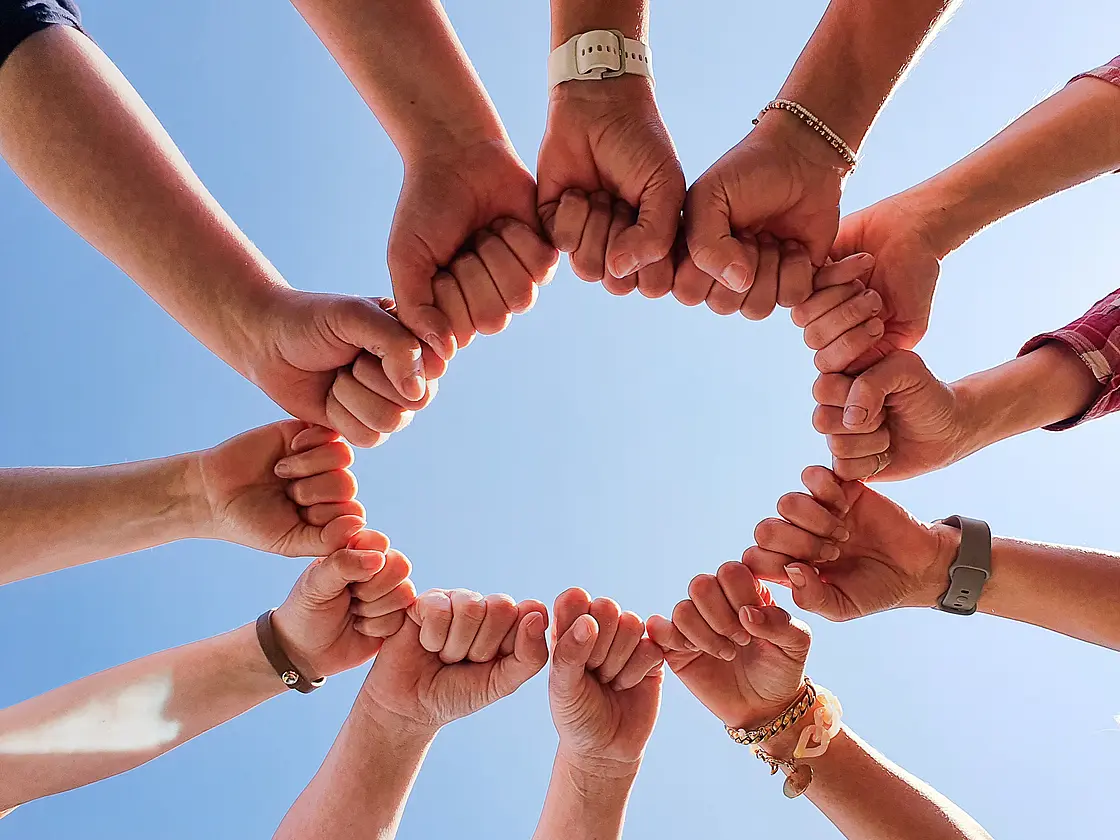 Mehrere ineinandergreifende Hände bilden einen Kreis vor blauem Himmel als Symbol für Selbsthilfe, Gemeinschaft und gegenseitige Unterstützung.