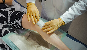 Mit Handschuhen versehene Hände legen einen Verband am Knie einer Patientin an.