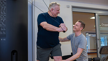 Ein Patient führt eine Übung an einem Trainingsgerät aus, während ein Therapeut unterstützend an seiner Seite steht.