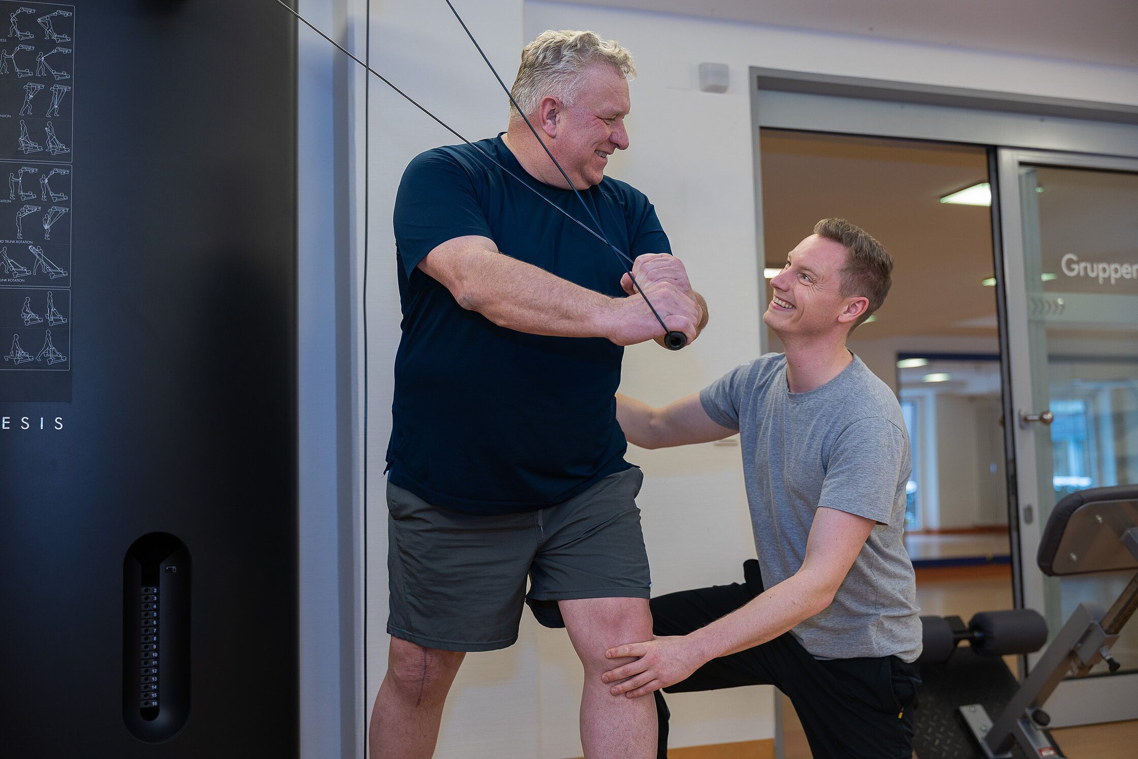 Ein Patient führt eine Übung an einem Trainingsgerät aus, während ein Therapeut unterstützend an seiner Seite steht.