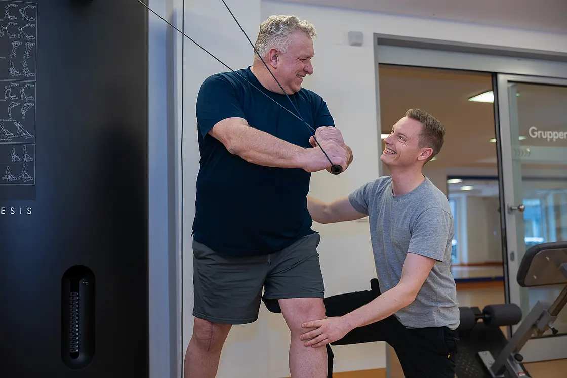 Ein Patient führt eine Übung an einem Trainingsgerät aus, während ein Therapeut unterstützend an seiner Seite steht.