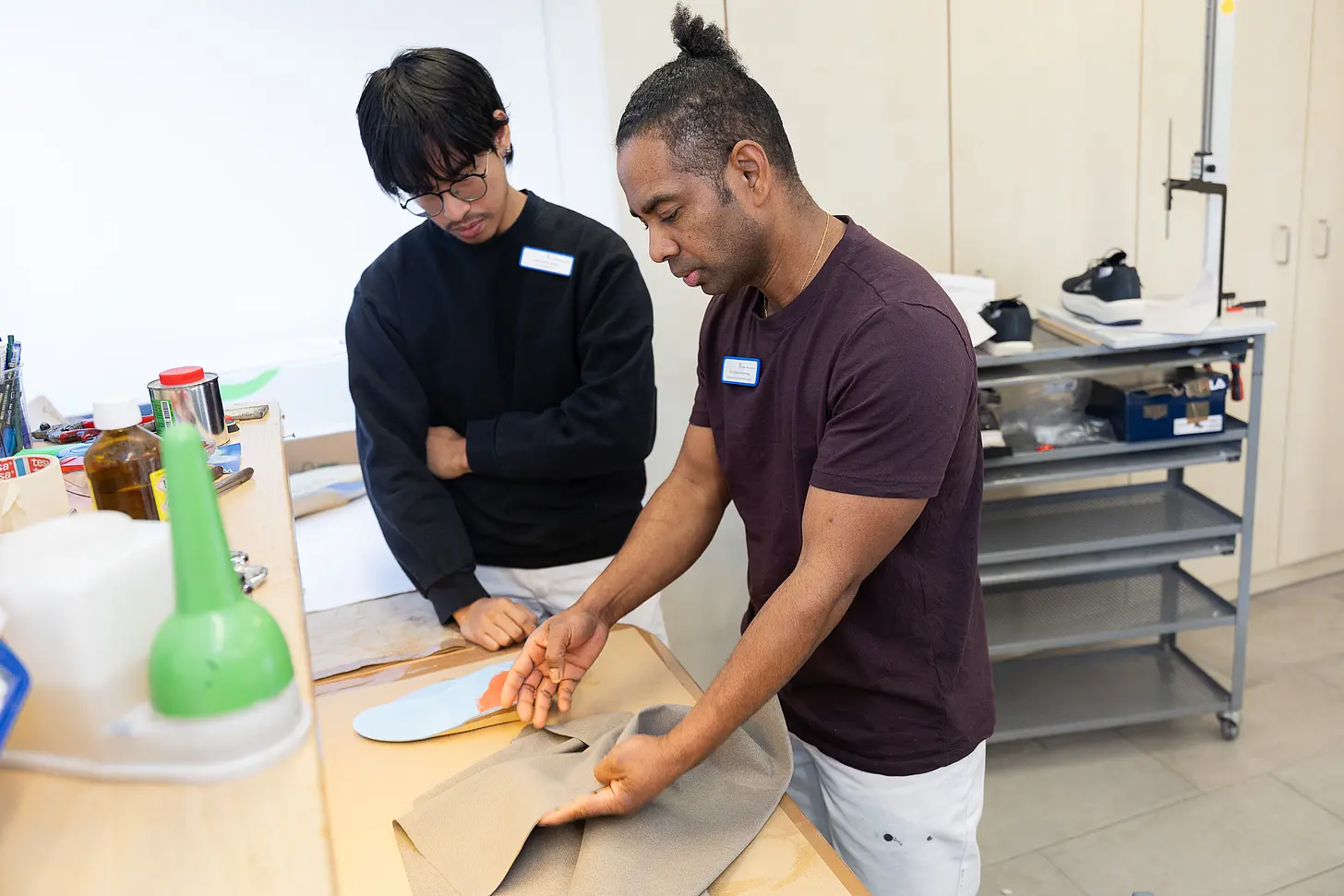 Ein Auszubildender und sein Praxisanleiter stehen an einer Werkbank in der orthopädischen Werkstatt und begutachten gemeinsam Materialien für eine orthopädische Schuheinlage.