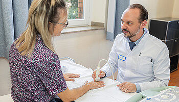 PD Dr. med. Peter Korsten untersucht die Hand einer sitzenden Patientin in einem Patientenzimmer.