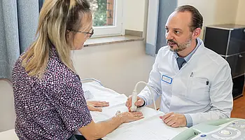 PD Dr. med. Peter Korsten untersucht die Hand einer sitzenden Patientin in einem Patientenzimmer.