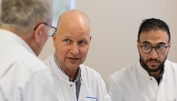 Ein Patient sitzt Chefarzt Dr. Frank Horst in weißem Kittel gegenüber und spricht mit ihm. Ein zweiter Arzt sitzt daneben und hört dem Gespräch zu.