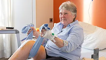 Eine ältere Frau sitzt im Krankenhausbett und hält ein Tuch, mit dem sie ihr angewinkeltes Bein unterstützt. An ihrem Arm ist ein medizinischer Zugang befestigt.