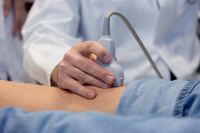 Ein Arzt führt einen Ultraschallkopf über das Knie eines Kindes; Kontaktgel ist auf der Haut sichtbar.