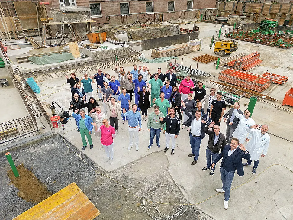 Mitarbeitende des St. Josef-Stifts stehen auf der Baustelle des OP- und Funktionsgebäudes und winken in die Kamera; im Hintergrund sind Bauarbeiten und Baumaterialien sichtbar.