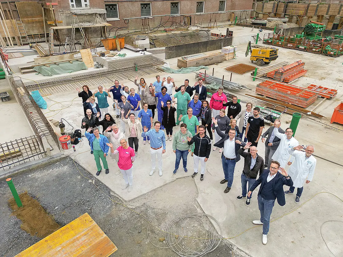 Mitarbeitende des St. Josef-Stifts stehen auf der Baustelle des OP- und Funktionsgebäudes und winken in die Kamera; im Hintergrund sind Bauarbeiten und Baumaterialien sichtbar.