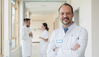 PD Dr. med. Peter Korsten steht mit verschränkten Armen auf einem Klinikflur, im Hintergrund sind Mitarbeitende zu sehen.