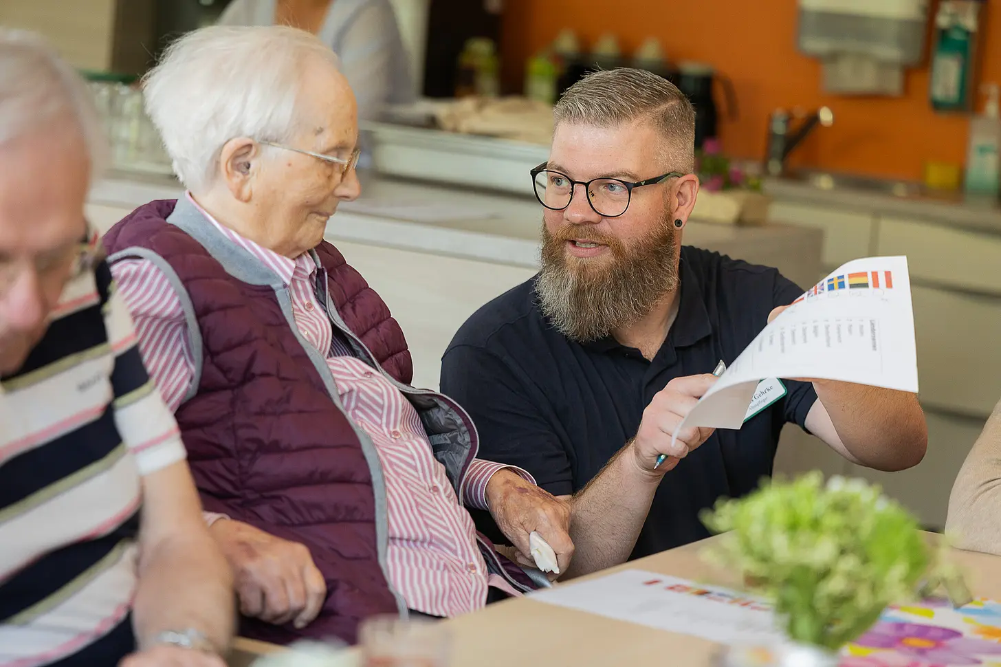 Mitarbeiter erklärt einer älteren Frau ein Dokument und zeigt auf eine Liste mit verschiedenen Flaggen, beide sitzen an einem Tisch.