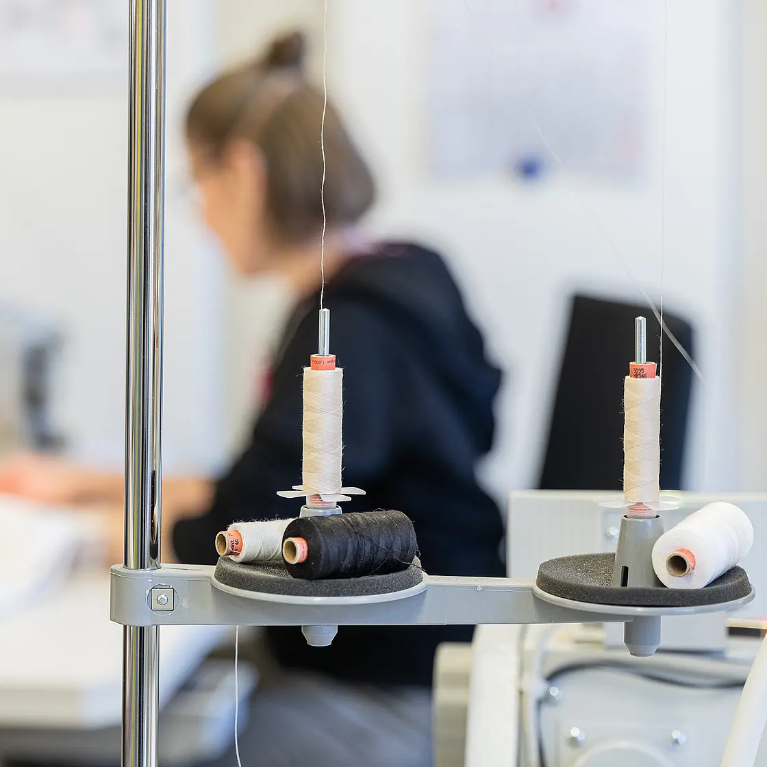 Blick auf einen Näharbeitsplatz mit Industrienähmaschine, Garnrollen und textilen Materialien.
