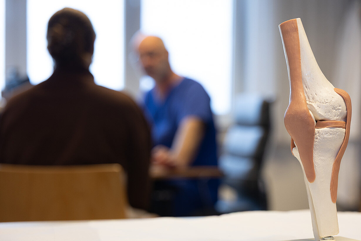 Knie-Modell im Vordergrund mit Arztgespräch im Hintergrund