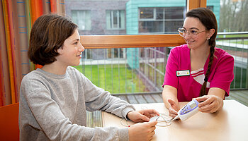 Pflegefachkraft erklärt einem Kind ein medizinisches Messgerät an einem Tisch am Fenster.