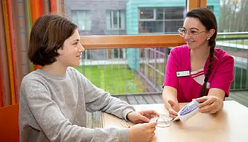 Pflegefachkraft erklärt einem Kind ein medizinisches Messgerät an einem Tisch am Fenster.