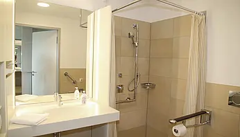 Ebenerdige Dusche mit Haltegriffen, Duschsitz und Vorhang, helle Fliesen und klare Linien.