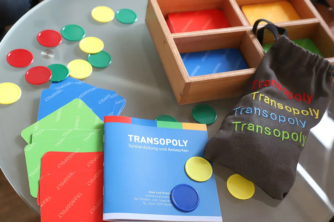 Spielmaterial „Transopoly“ mit Karten, Spielsteinen und Anleitung liegt auf einem Tisch.