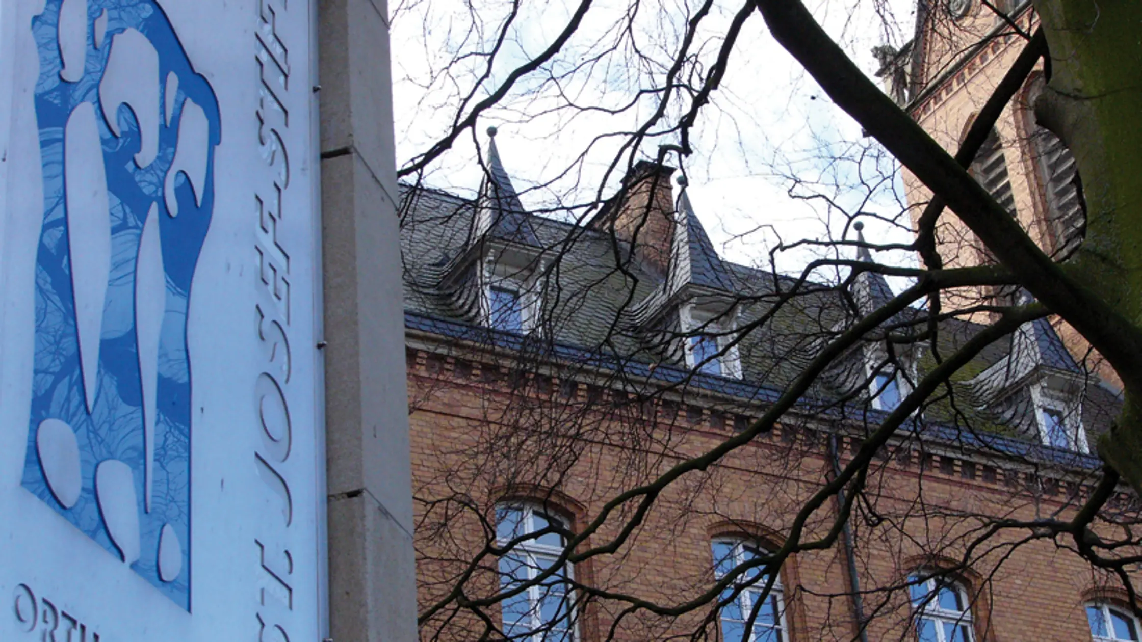 Außenansicht eines Backsteingebäudes des St. Josef-Stift Sendenhorst mit Schild des Orthopädischen Zentrums, teilweise von Bäumen verdeckt.