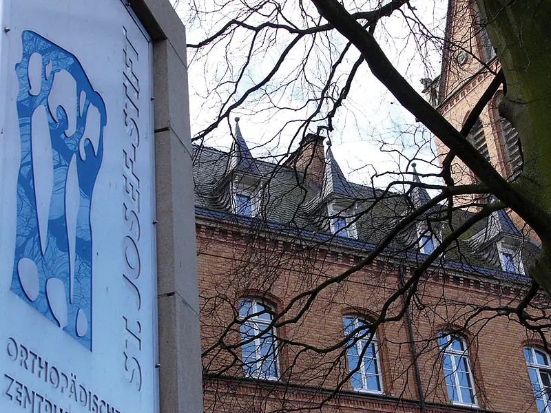 Außenansicht eines Backsteingebäudes des St. Josef-Stift Sendenhorst mit Schild des Orthopädischen Zentrums, teilweise von Bäumen verdeckt.