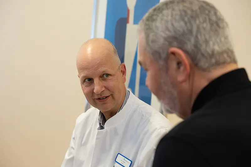 Chefarzt Dr. Frank Horst in weißem Kittel spricht mit einem Patienten. Beide sitzen sich gegenüber und schauen sich an.
