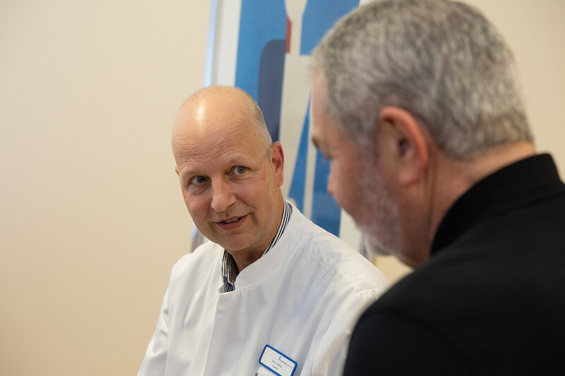 Chefarzt Dr. Frank Horst in weißem Kittel spricht mit einem Patienten. Beide sitzen sich gegenüber und schauen sich an.