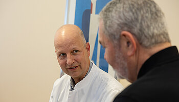 Ein Arzt in weißem Kittel spricht mit einem Patienten. Beide sitzen sich gegenüber und schauen sich an.