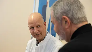 Ein Arzt in weißem Kittel spricht mit einem Patienten. Beide sitzen sich gegenüber und schauen sich an.