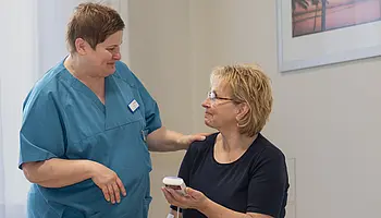 Eine Pflegefachperson erklärt einer Patientin die Anwendung eines TENS-Geräts und legt dabei beruhigend die Hand auf ihre Schulter.