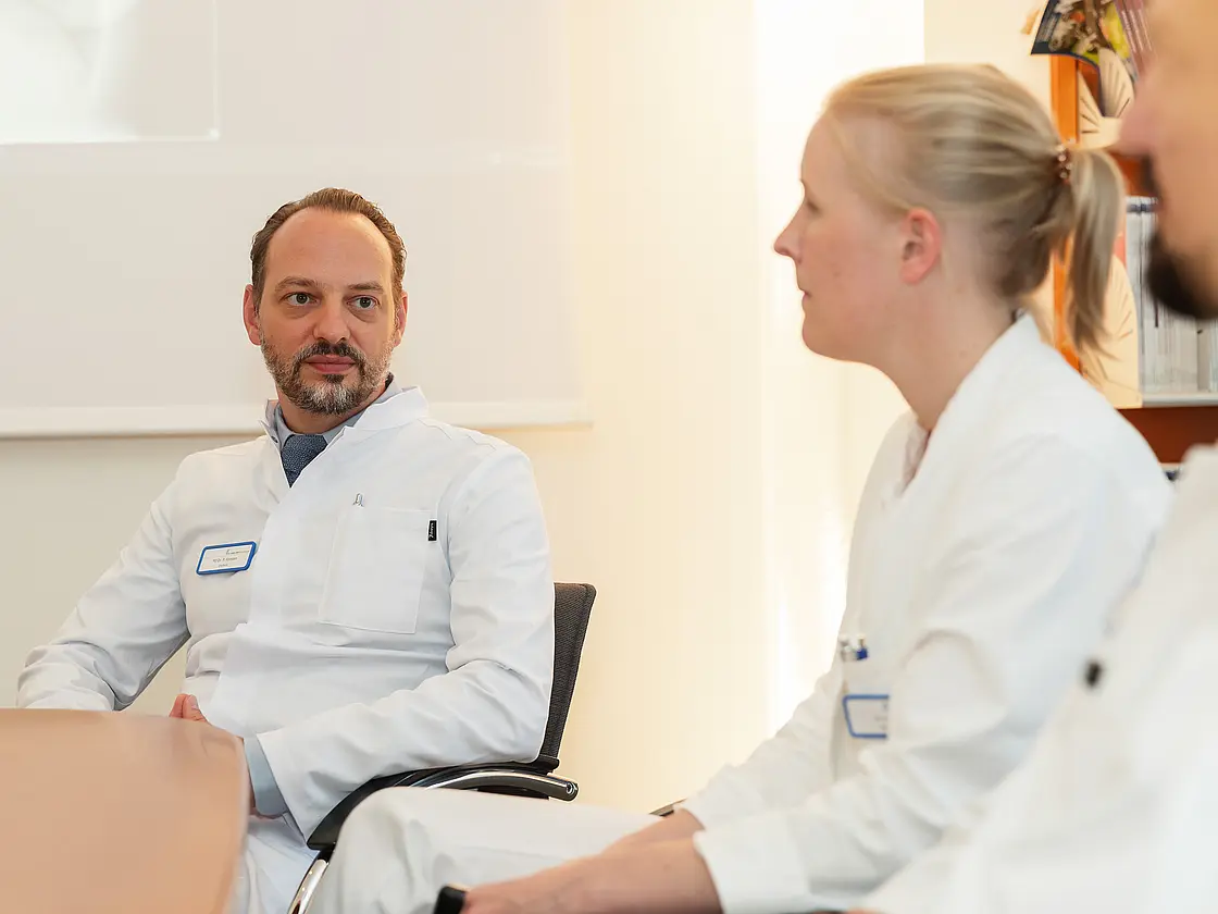 PD Dr. med. Peter Korsten sitzt an einem Konferenztisch und hört einer Kollegin zu.