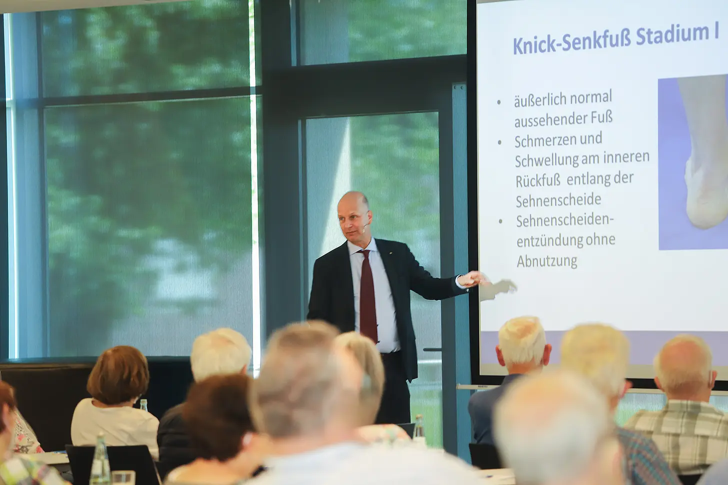 Chefarzt Dr. Frank Horst steht im Anzug vor einer Leinwand und hält einen Vortrag. Auf der Präsentation ist der Titel „Knick-Senkfuß Stadium I“ zu lesen. Vor ihm sitzen Zuhörerinnen und Zuhörer an Tischen und verfolgen den Vortrag.