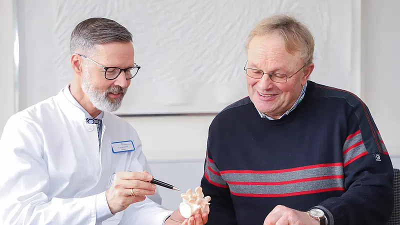 Chefarzt Dr. Christian Brinkmann erklärt einem Patienten anhand eines Wirbelsäulenmodells einen medizinischen Befund.