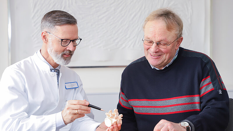 Chefarzt Dr. Christian Brinkmann erklärt einem Patienten anhand eines Wirbelsäulenmodells einen medizinischen Befund.