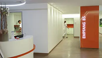 Stationsflur mit Empfangstresen und orangefarbener Wandbeschriftung „Empfang B5“, indirekte Beleuchtung entlang der Wände.