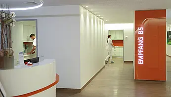 Stationsflur mit Empfangstresen und orangefarbener Wandbeschriftung „Empfang B5“, indirekte Beleuchtung entlang der Wände.