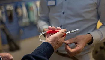 Nahaufnahme einer Hand, die eine rote Schere hält, während eine zweite Person auf Details des Hilfsmittels zeigt.