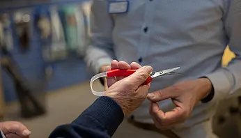 Nahaufnahme einer Hand, die eine rote Schere hält, während eine zweite Person auf Details des Hilfsmittels zeigt.