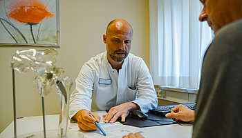Ein Arzt erläutert einem sitzenden Patienten anhand eines Hüftgelenkmodells und schriftlicher Unterlagen eine geplante Behandlung.