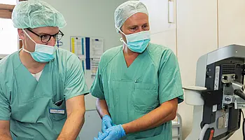 Chefarzt Dr. med. Matthias Boschin und ein weiterer Mitarbeiter in OP-Kleidung stehen an einem medizinischen Gerät und stimmen Abläufe ab.